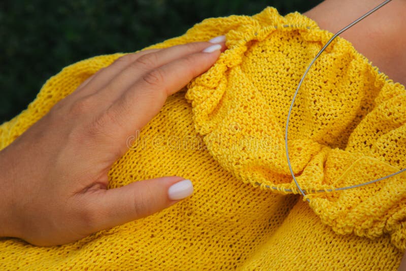 A young girl with long blond hair knits a yellow sweater in the garden in the summer. woman makes clothes with hands closeup. A young girl with long blond hair knits a yellow sweater in the garden in the summer. woman makes clothes with hands closeup.