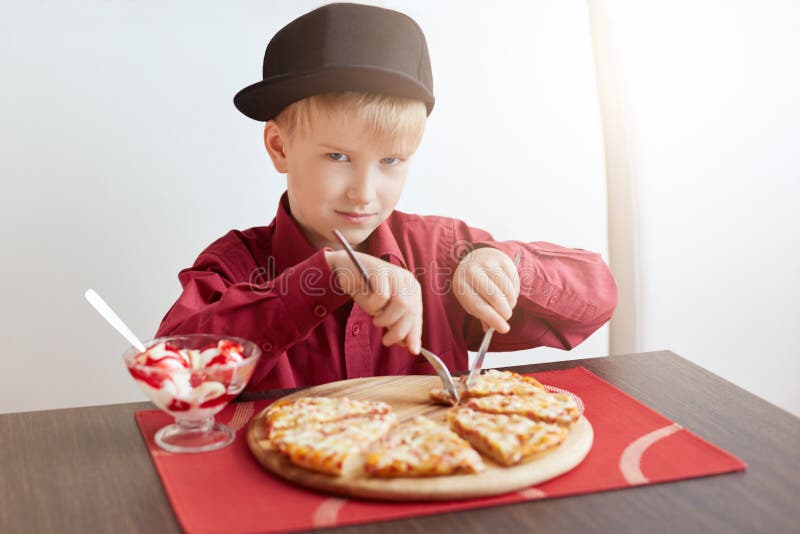 Ein kleiner blonder Junge mit den blauen Augen, die rotes Hemd tragen und die stilvolle Kappe, die im angenehmen Café köstliche P lizenzfreie stockfotos Ein kleiner blonder Junge mit den blauen Augen, die rotes Hemd tragen und die stilvolle Kappe, die im angenehmen Café köstliche P lizenzfreie stockfotos