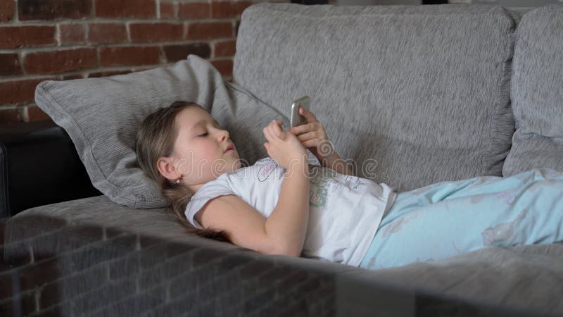Ein Kleines Mädchen Liegt Zu Hause Auf Der Couch Und Schaut in Ein Smartphone. Das Kind Spielt Und Sieht Das Video Stock Footage - Video von menschlich, telefon: 215207584