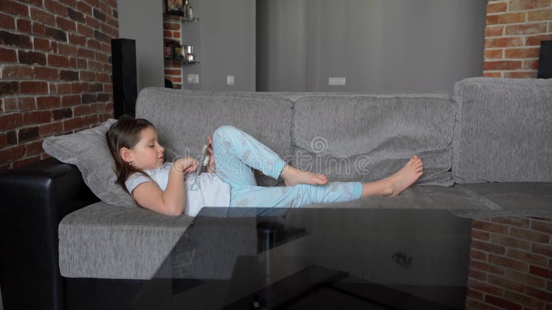 Ein Kleines Mädchen Liegt Zu Hause Auf Der Couch Und Schaut in Ein Smartphone. Das Kind Spielt Und Sieht Das Video Stock Footage - Video von menschlich, relax: 215209504