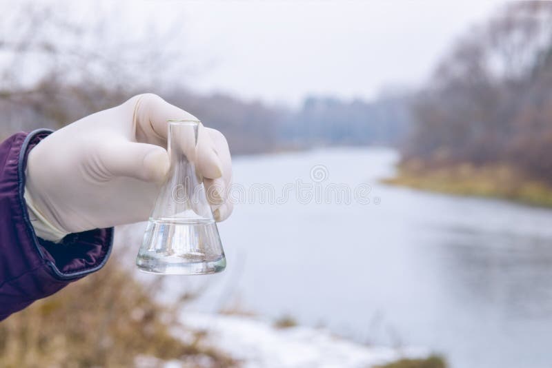 A laboratorian is holding a test tube with transparent river water sample inside at the river bank background. A laboratorian is holding a test tube with transparent river water sample inside at the river bank background