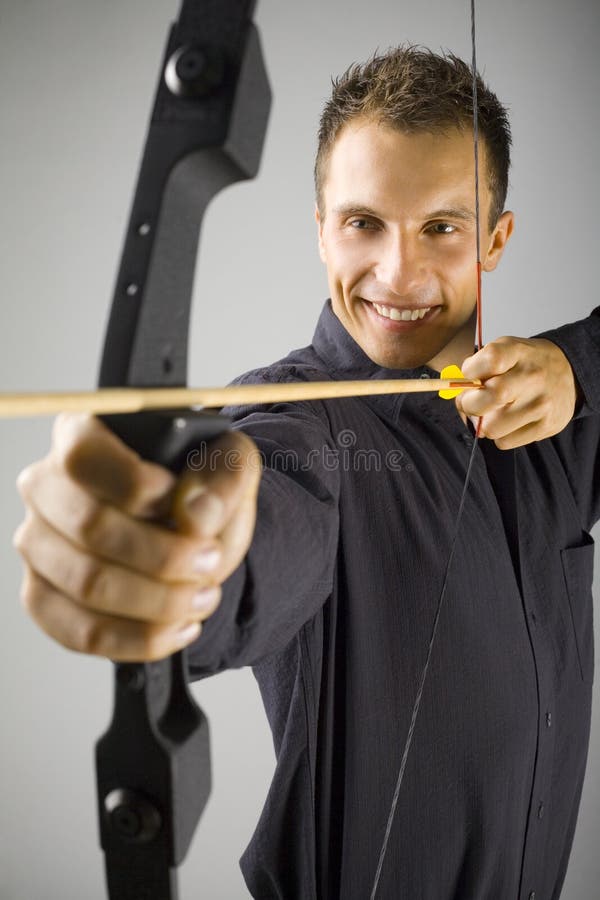 Young, elegant man in black shirt holding bow. Smiling and shooting to target. Gray background, front view. Young, elegant man in black shirt holding bow. Smiling and shooting to target. Gray background, front view