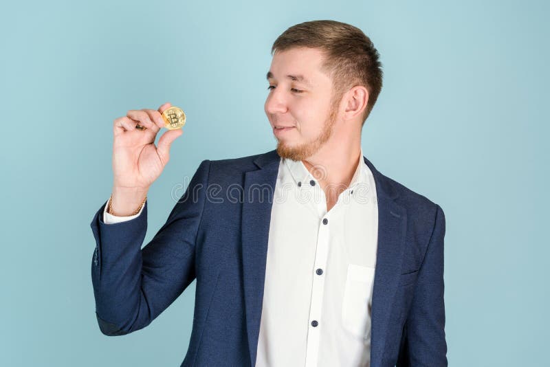 Ein lächelnder bärtiger Hipster-Geschäftsmann betrachtet die Bitcoin-Goldmünze in seiner Hand lizenzfreie stockbilder