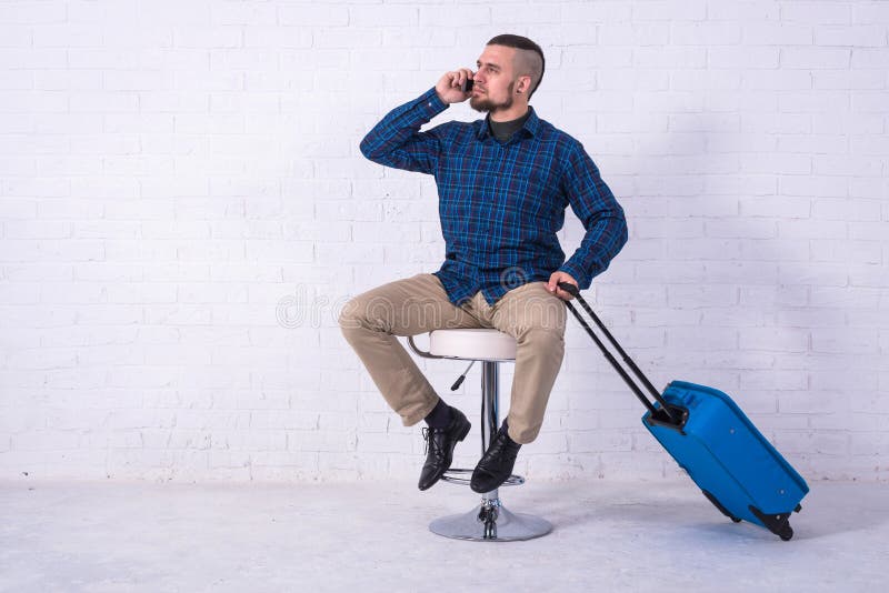 Ein Mann mit einem blauen Koffer sitzt auf einem Stuhl nahe einer wei?en Backsteinmauer und spricht am Telefon lizenzfreies stockbild Ein Mann mit einem blauen Koffer sitzt auf einem Stuhl nahe einer wei?en Backsteinmauer und spricht am Telefon lizenzfreies stockbild