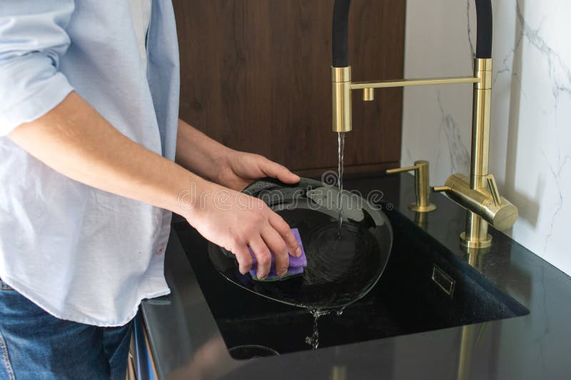 A man washes a black plate in the kitchen in the sink under water. A man washes a black plate in the kitchen in the sink under water