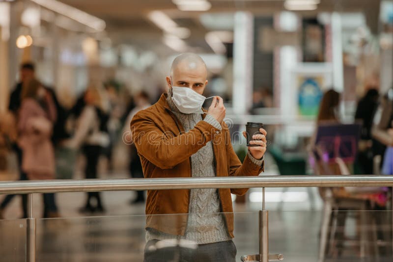 Ein Mann zieht eine Maske an, während er im Einkaufszentrum eine Tasse Kaffee hält stockbild