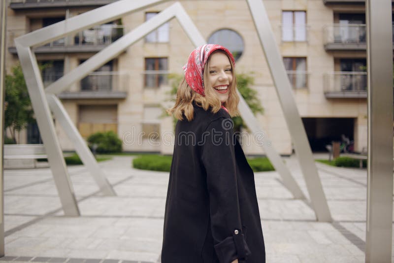 A girl in a business suit with a red scarf on her head against the background of a building, turns to the camera and laughs. Fashion photo of a blonde model poses, in a suit and with a red scarf. A girl in a business suit with a red scarf on her head against the background of a building, turns to the camera and laughs. Fashion photo of a blonde model poses, in a suit and with a red scarf