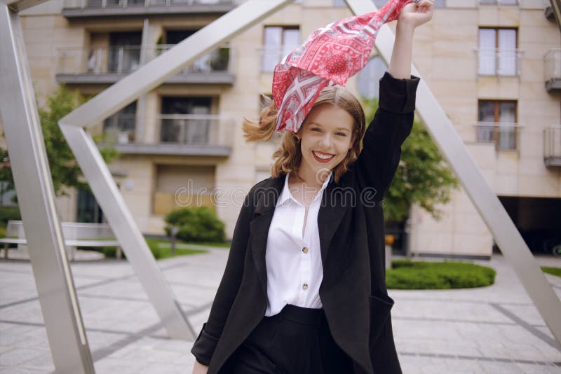 A girl in a business suit with a red scarf on her head against the background of a building, turns to the camera and laughs. Fashion photo of a blonde model poses, in a suit and with a red scarf. A girl in a business suit with a red scarf on her head against the background of a building, turns to the camera and laughs. Fashion photo of a blonde model poses, in a suit and with a red scarf