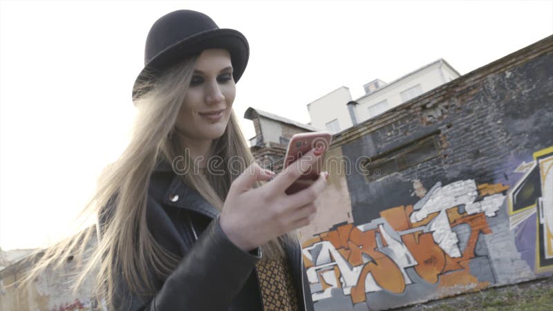 Ein Mädchen im schwarzen Hut und in der Lederjacke gehend hinunter die Straße gegen die Wand bedeckt durch graffity, Schmutzart stockfotografie Ein Mädchen im schwarzen Hut und in der Lederjacke gehend hinunter die Straße gegen die Wand bedeckt durch graffity, Schmutzart stockfotografie