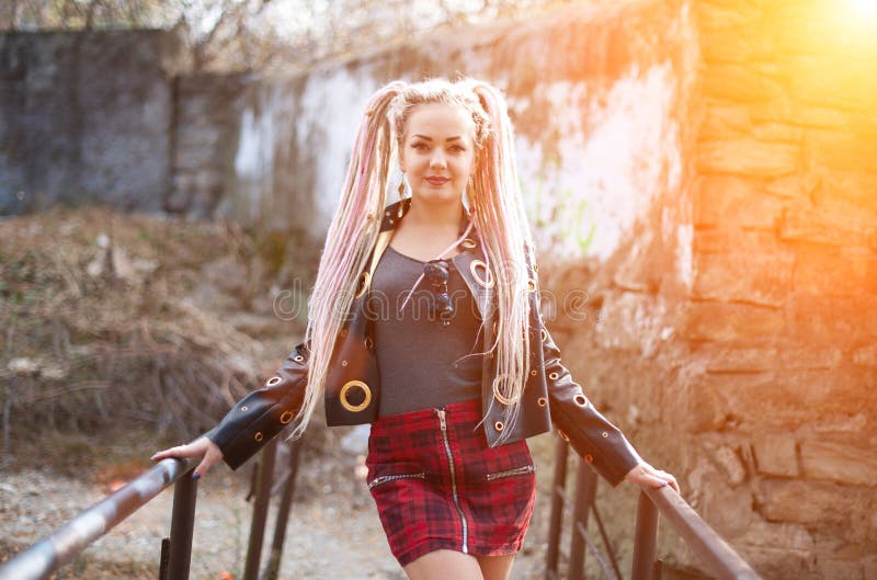 Ein Mädchen mit Dreadlocks in einer Lederjacke und in einem kurzen Rock steht vor dem hintergrund einer alten Steinwand in den St lizenzfreies stockfoto Ein Mädchen mit Dreadlocks in einer Lederjacke und in einem kurzen Rock steht vor dem hintergrund einer alten Steinwand in den St lizenzfreies stockfoto