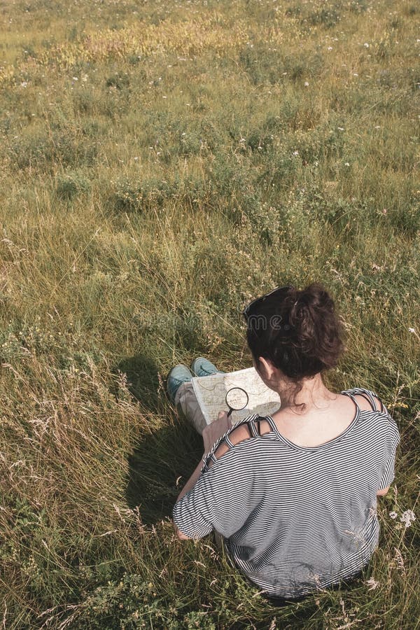Ein Mädchentourist in einer gestreiften Strickjacke sitzt auf einem Gebiet unter dem Gras und betrachtet eine Karte durch ein Ver stockfotos Ein Mädchentourist in einer gestreiften Strickjacke sitzt auf einem Gebiet unter dem Gras und betrachtet eine Karte durch ein Ver stockfotos