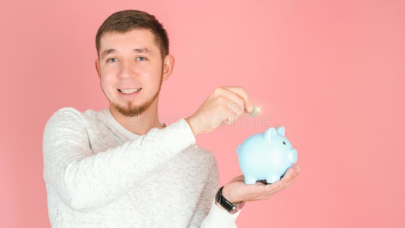Ein männlicher Investor legt eine Münze in einem blauen Sparschwein auf einem rosa Hintergrund stockbild