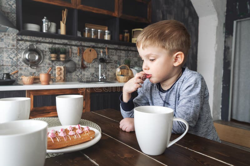 Ein nachdenklicher kleiner Junge sitzt an einem Tisch mit einem circley Tee und Kuchen Lecken seines Fingers Porträt stockbilder Ein nachdenklicher kleiner Junge sitzt an einem Tisch mit einem circley Tee und Kuchen Lecken seines Fingers Porträt stockbilder
