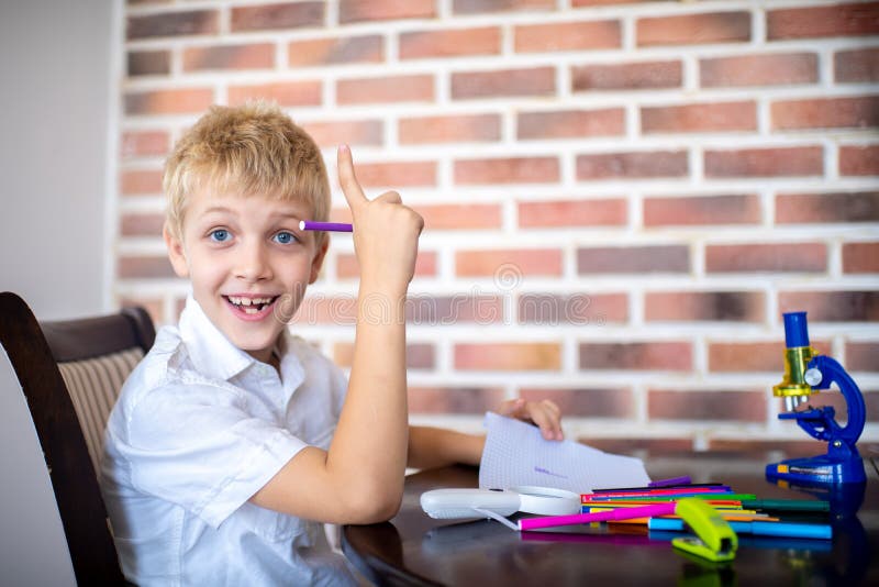 Ein netter Junge fand eine Idee. Glücklicher netter Junge fand eine Idee Junger Schülerstudent sitzt in einem Raum nahe einer Backsteinmauer, ist auf dem Tisch zerstreute Markierungen und Büroartikel Schulsystemlebensstil von neuen Leuten