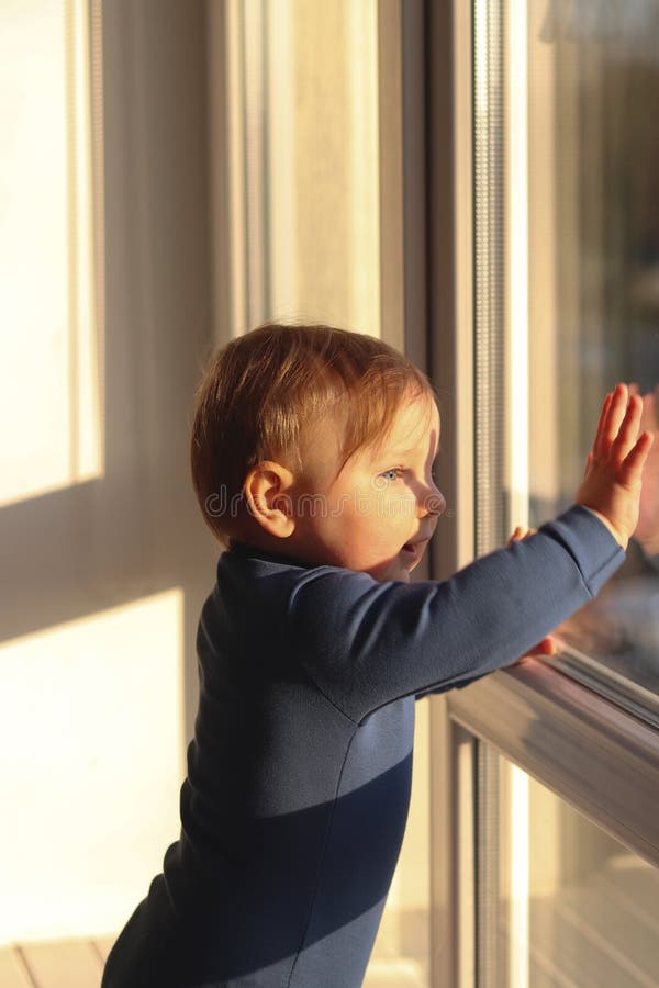 Ein niedlicher kleiner Junge steht am Fenster und blickt auf die Straße. Sonnenaufgang oder Sonnenuntergang stockfotos