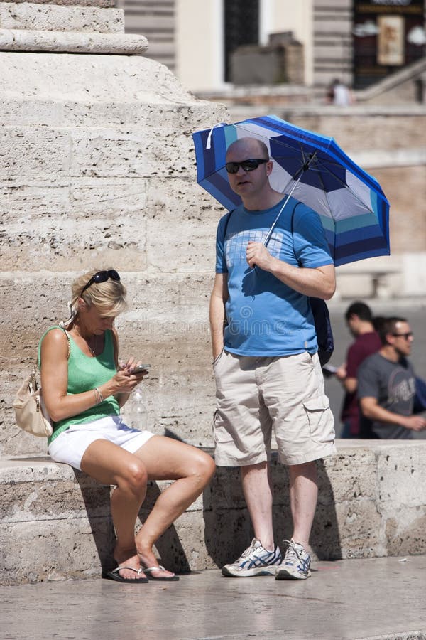 Two tourists, a couple, they are the fountain of Piazza del Popolo in Rome (Italy). The man is standing and protects himself from the sun with an umbrella in blue stripes. The woman is sitting and checking her cell phone. Two tourists, a couple, they are the fountain of Piazza del Popolo in Rome (Italy). The man is standing and protects himself from the sun with an umbrella in blue stripes. The woman is sitting and checking her cell phone.