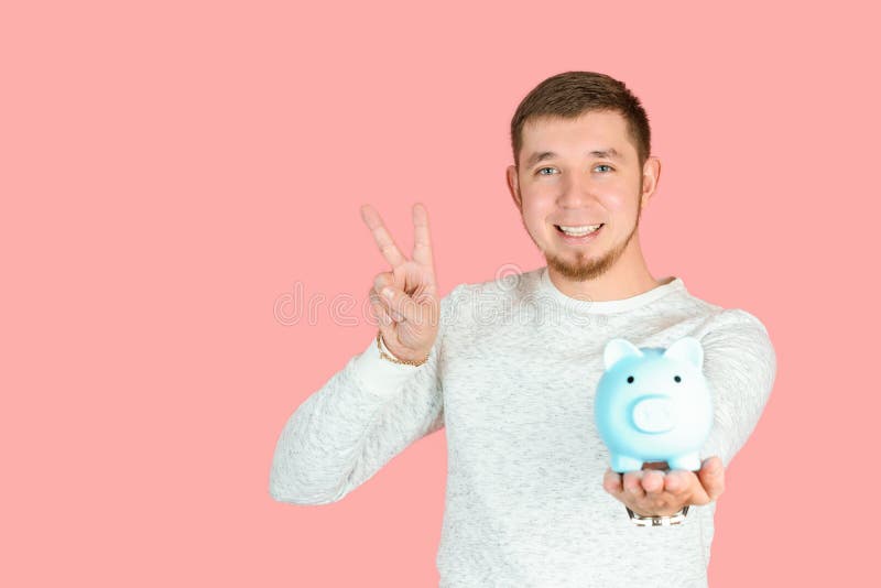 Ein rot gebärdeter Mann in einem weißen Pullover hält ein blaues Sparschwein in der Hand und zeigt eine Siegesgeste Copyspace stockbilder