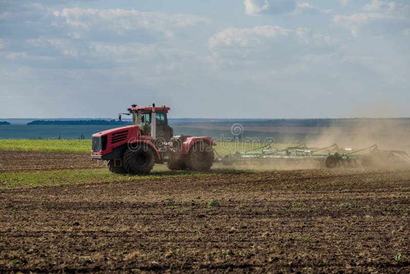 A red farm tractor in a cloud of dust cultivates the soil in the field with a cultivator after harvest. Summer sunny day. Fertile land. Modern agricultural machinery. Copy space. High quality photo. A red farm tractor in a cloud of dust cultivates the soil in the field with a cultivator after harvest. Summer sunny day. Fertile land. Modern agricultural machinery. Copy space. High quality photo