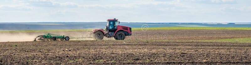 A red farm tractor in a cloud of dust cultivates the soil in the field with a cultivator after harvest. Summer sunny day. Fertile land. Modern agricultural machinery. Copy space. Banner. A red farm tractor in a cloud of dust cultivates the soil in the field with a cultivator after harvest. Summer sunny day. Fertile land. Modern agricultural machinery. Copy space. Banner.