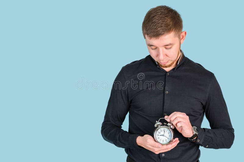 Ein rötlich bärtiger Mann hält einen Wecker in den Händen und schaut hinab, während er auf einem blauen Hintergrund steht stockbilder