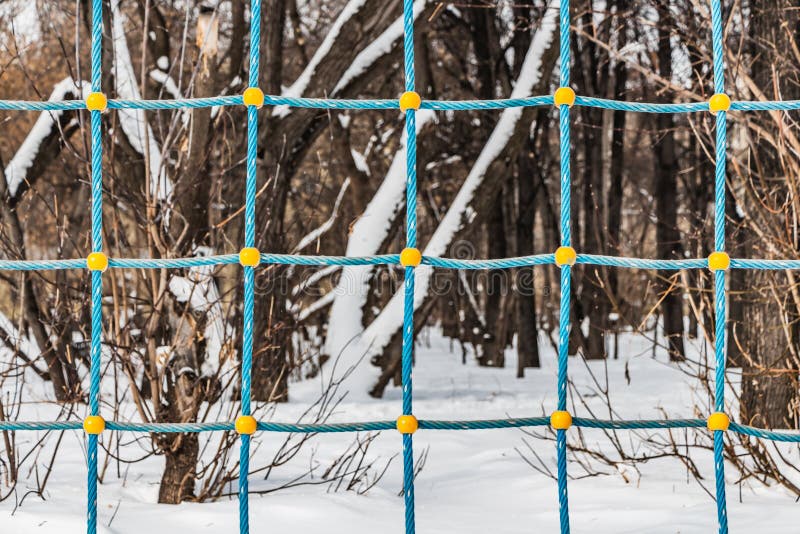 Ein schneebedeckter Park mit braunen Bäumen durch ein blaues und gelbes Seilnetz von einem Spielplatz der Kinder im Winter stockbild Ein schneebedeckter Park mit braunen Bäumen durch ein blaues und gelbes Seilnetz von einem Spielplatz der Kinder im Winter stockbild