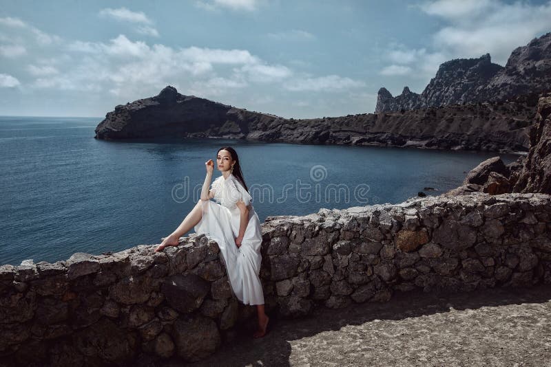 Ein schönes Mädchen, Braut, in einem weißen Kleid sitzt einen Steinzaun auf einer Klippe vor dem Hintergrund des Ozeans und der B lizenzfreies stockbild