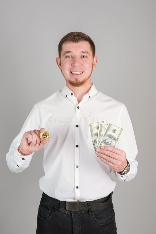 Ein selbstbewusster Geschäftsmann in einem weißen Hemd hält eine Bitcoin-Goldmünze und einen Fan von Papier-Dollar-Scheinen in sei lizenzfreies stockbild