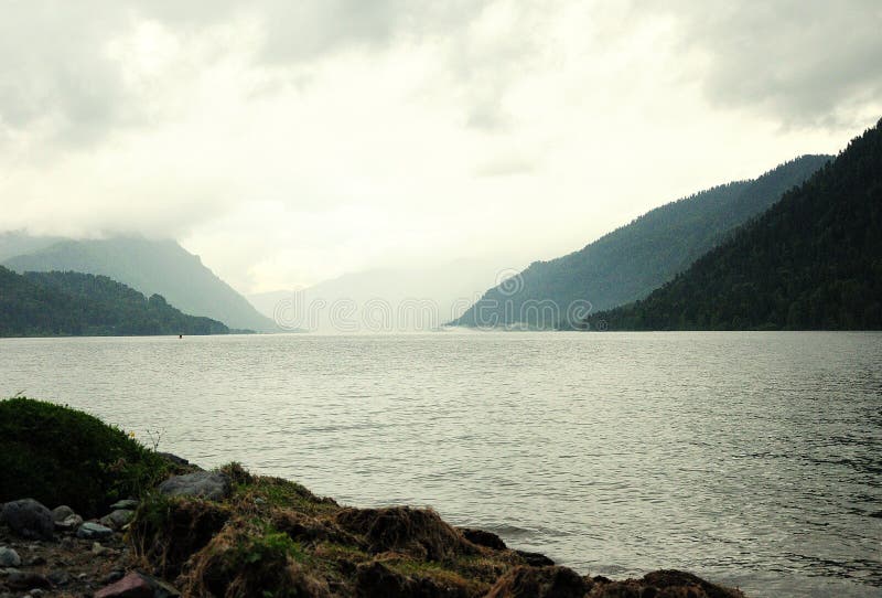 Ein Teil des felsigen Ufers eines großen Bergsees bei bewölktem Wetter mit Wolken auf den Gipfeln lizenzfreie stockfotos