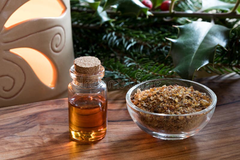 A bottle of myrrh essential oil with myrrh resin in a glass bottle, and spruce branches, holly branches and an aroma lamp in the background. A bottle of myrrh essential oil with myrrh resin in a glass bottle, and spruce branches, holly branches and an aroma lamp in the background
