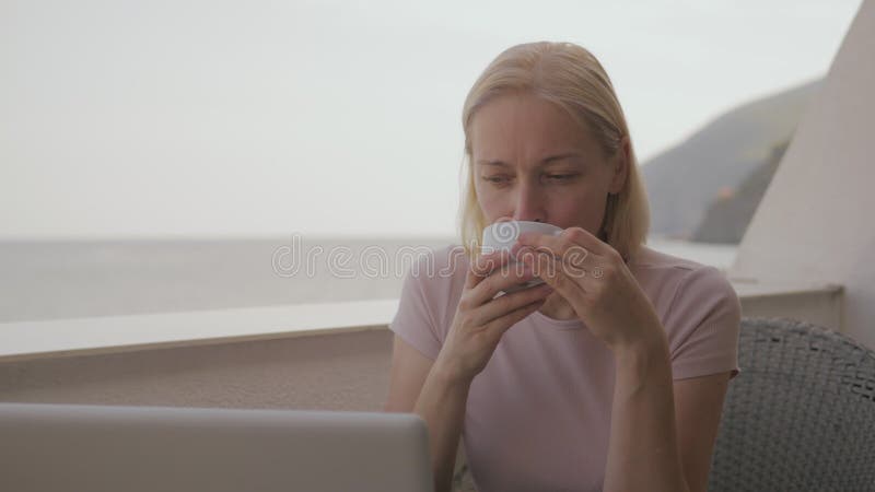 Eine Frau arbeitet auf einem Laptop auf dem Balkon eines Hotelzimmers und trinkt an einem sonnigen Sommertag Kaffee.