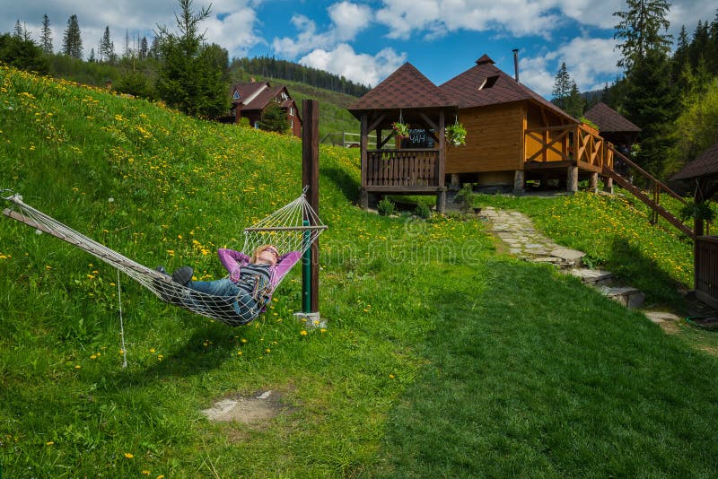 Eine Frau, die in der Hängematte in den Bergen stillsteht Holzhäuser auf dem Hintergrund und dem grünen Gras lizenzfreies stockfoto Eine Frau, die in der Hängematte in den Bergen stillsteht Holzhäuser auf dem Hintergrund und dem grünen Gras lizenzfreies stockfoto
