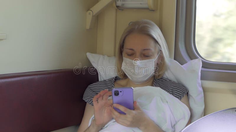 Eine Frau in einer medizinischen Schutzmaske liegt mit einem Mobiltelefon in einem Fahrgastraum.