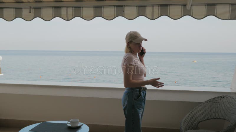 Eine Frau mit Baseballmütze, die auf dem Balkon eines Hotelzimmers mit Blick auf das Meer telefoniert.