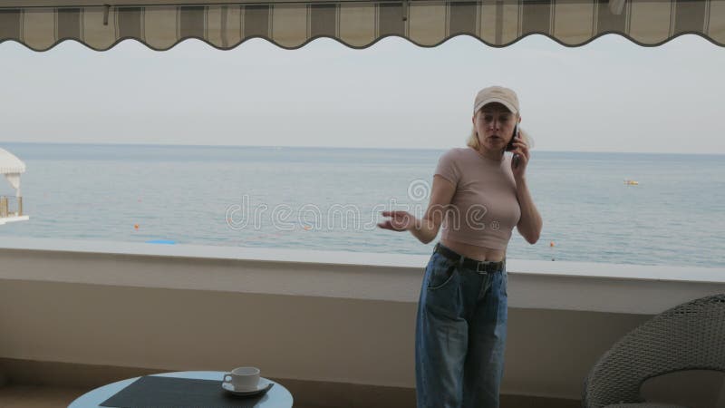 Eine Frau mit Baseballmütze, die nervös telefoniert, auf dem Balkon eines Hotelzimmers mit Blick auf das Meer.