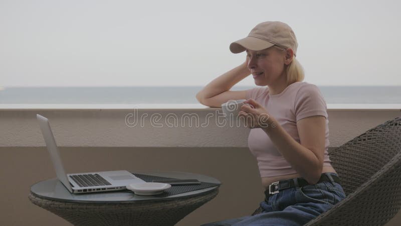 Eine Frau telefoniert und redet online auf einem Laptop auf einem Hotelbalkon mit Meerblick