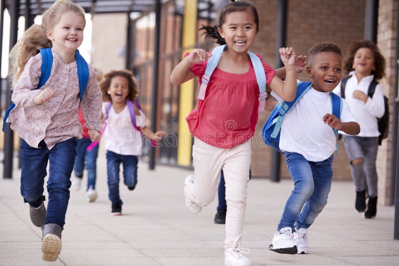 Eine Gruppe lächelnde multiethnische Schulkinder, die oben in einen Gehweg außerhalb ihres Säuglingsschulgebäudees eine Lektion stockfotografie