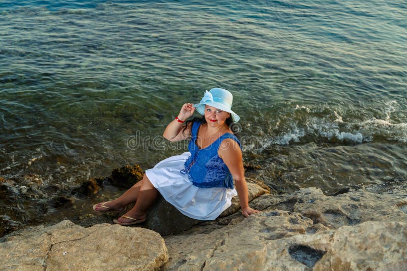 Eine junge Frau in einem weißen Rock und ein Sonnenmütze am Meer liegen auf einem Stein, der die Natur bewundert. lizenzfreie stockfotografie
