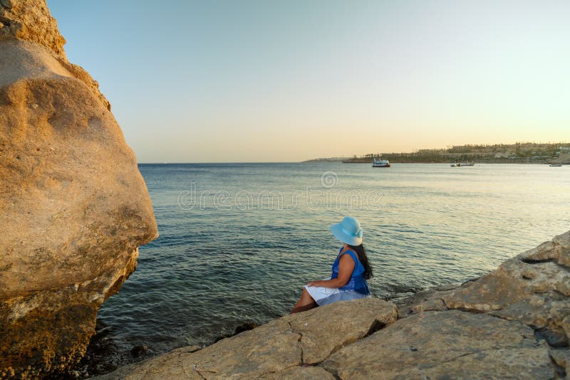Eine junge Frau in einem weißen Rock und ein Sonnenmütze am Meer sitzt auf einem Stein, der die Natur mit ihrem Rücken zur Kamera lizenzfreie stockbilder