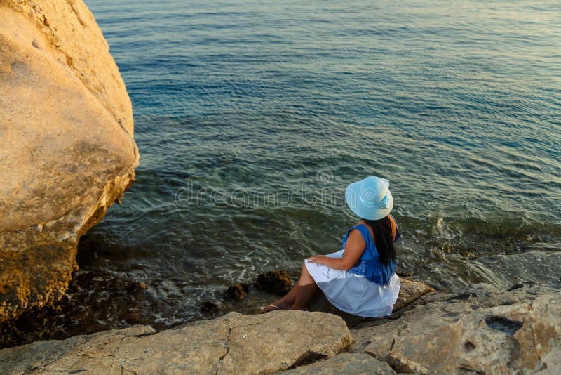 Eine junge Frau in einem weißen Rock und ein Sonnenmütze am Meer sitzt auf einem Stein, der die Natur mit ihrem Rücken zur Kamera stockbilder