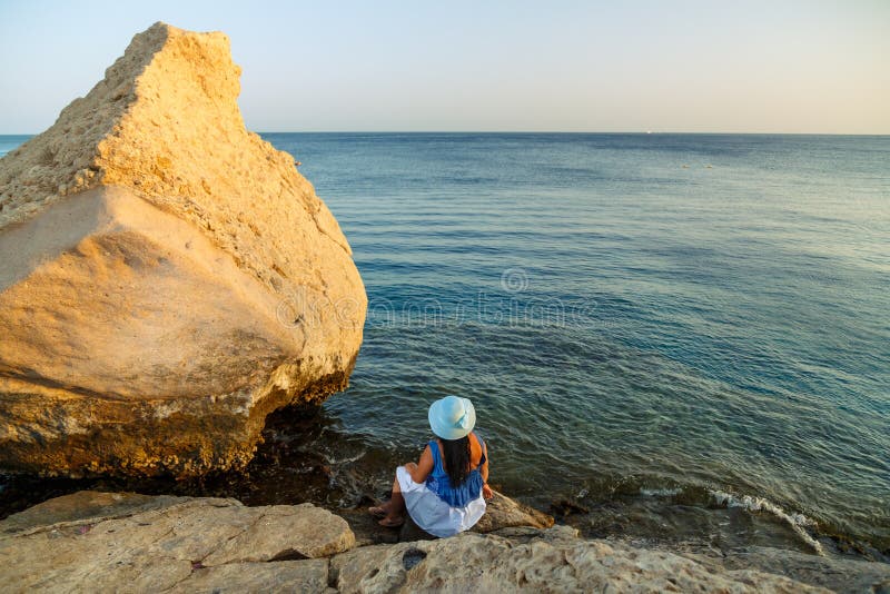 Eine junge Frau in einem weißen Rock und ein Sonnenmütze am Meer sitzt auf einem Stein, der die Natur mit ihrem Rücken zur Kamera lizenzfreie stockfotos