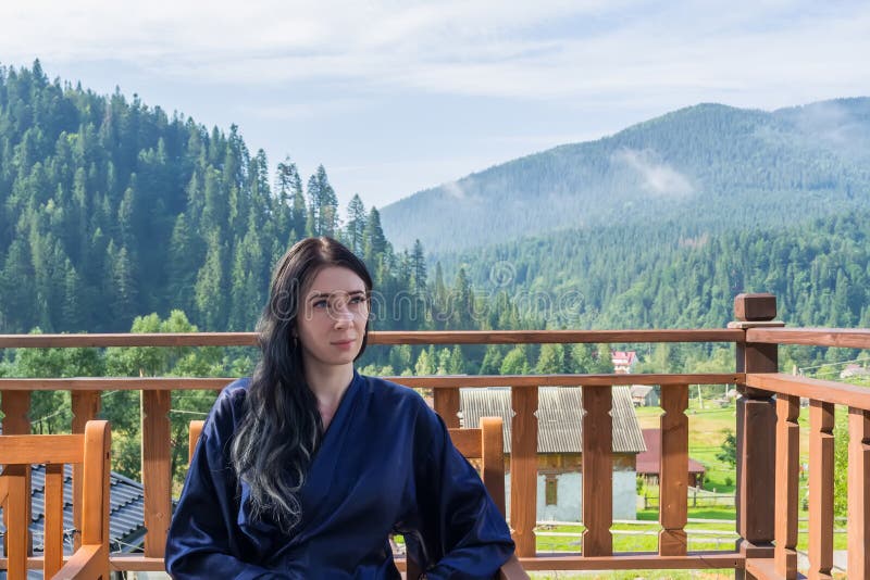 Eine junge Frau genießt eine Berglandschaft und sitzt in einem Bademantel auf dem Balkon stockbild Eine junge Frau genießt eine Berglandschaft und sitzt in einem Bademantel auf dem Balkon stockbild