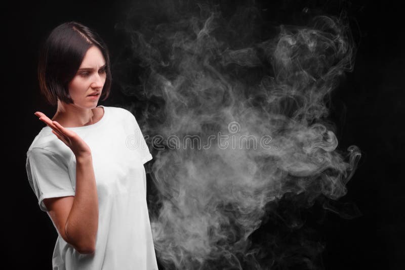 Eine junge Frau mag nicht den Rauch einer Zigarette oder der elektronischen Zigarette auf einem schwarzen Hintergrund Apfel- und lizenzfreies stockfoto Eine junge Frau mag nicht den Rauch einer Zigarette oder der elektronischen Zigarette auf einem schwarzen Hintergrund Apfel- und lizenzfreies stockfoto