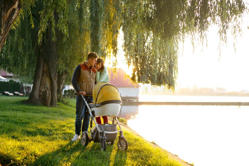 Eine junge liebevolle Familie geht durch den See mit einem Spaziergänger Das Lächeln erzieht Paare mit Baby Pram im Herbstpark Li stockbild Eine junge liebevolle Familie geht durch den See mit einem Spaziergänger Das Lächeln erzieht Paare mit Baby Pram im Herbstpark Li stockbild