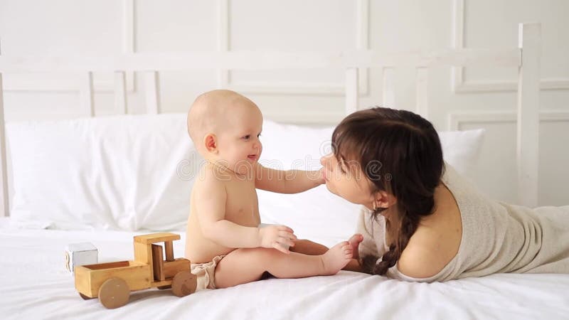 Eine junge Mutter spielt mit einem Kind in Windeln mit Holzspielzeug auf einem hellen Bett zu Hause und lächelt