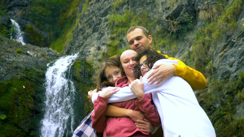 Eine kahlköpfige Frau, ihre Kinder und ihr Mann umarmten den Hintergrund eines Wasserfalls und schauen sich die Kamera an