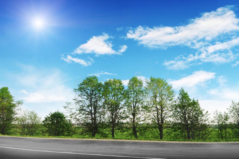 Eine leere Landstrasse mit Bäumen und Sträuchen an einem blauen Himmel mit Wolken und Sonne lizenzfreie stockfotografie