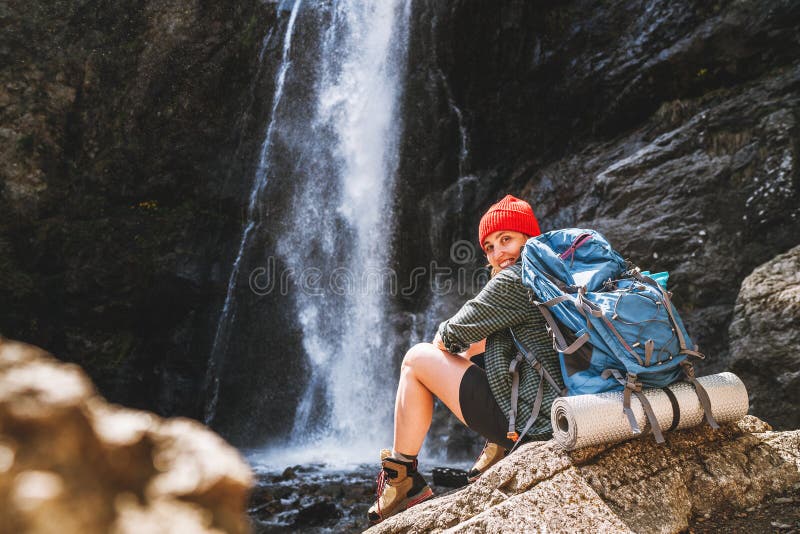 Eine lächelnde Frau mit einem Rucksack in rotem Hut in aktiven Trekkingbekleidung und Stiefeln in der Nähe des Wasserfalles des Ge lizenzfreie stockfotografie