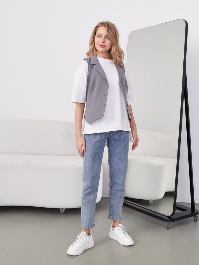 Eine lächelnde Frau steht in einem modernen Raum mit Sofa und großem Spiegel. Sie trägt eine graue westliche blaue Jeans und lizenzfreies stockfoto