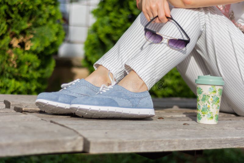 Eine moderne Frau mit gestreiften Hosen und blauen den Turnschuhen, die auf einer Bank im Park sitzen und Sonnenbrille, Kaffeetas stockfotografie Eine moderne Frau mit gestreiften Hosen und blauen den Turnschuhen, die auf einer Bank im Park sitzen und Sonnenbrille, Kaffeetas stockfotografie