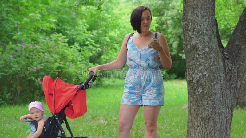 Eine Mutter rüttelt ein Kind in einem Spaziergänger und schreibt nervös eine Mitteilung in ihren Handy